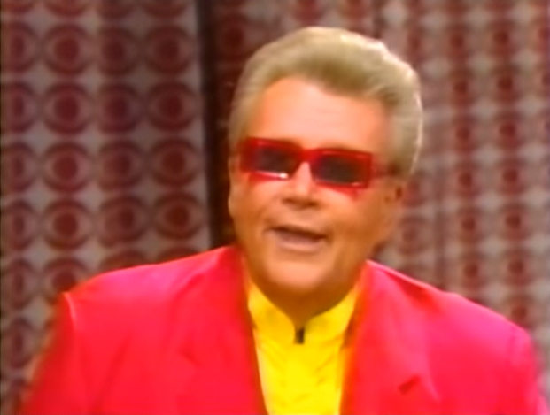Rod is wearing a salmon-colored jacket, yellow collarless silk shirt & red rectangular sunglasses