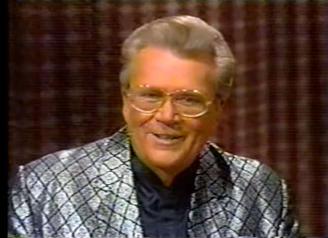 Rod is wearing a shiny silver-checked jacket & dark-blue collarless silk shirt