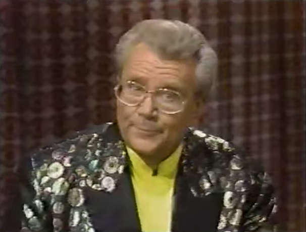 Rod is wearing a black jacket with silver dots, black lapels & a yellow collarless silk shirt