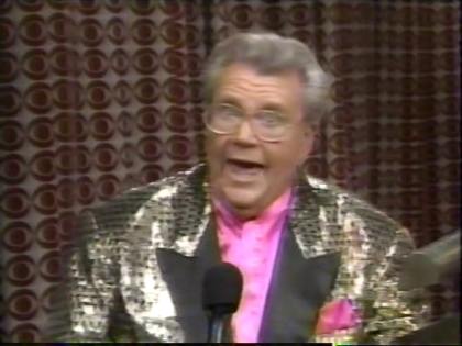 Rod is wearing a gold jacket with black squares and black lapels & a fuchsia collarless silk shirt with matching pocket square