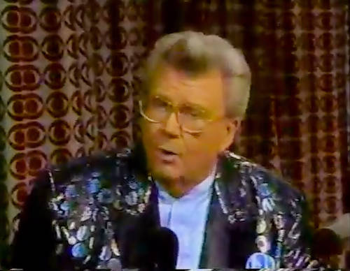 Rod is wearing a black jacket w/shiny circles  black lapels & powder-blue collarless silk shirt w/matching pocketsquare