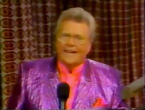 Rod is wearing a shiny purple/purple striped jacket & red collarless silk shirt w/matching pocket square