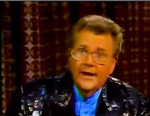 Rod is wearing a black jacket with shiny dots & blue collarless silk shirt