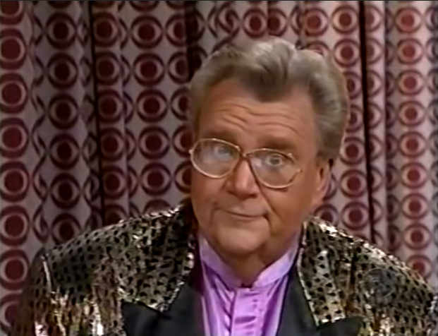 Rod is wearing a gold jacket with black spots & black lapels & a violet collarless silk shirt