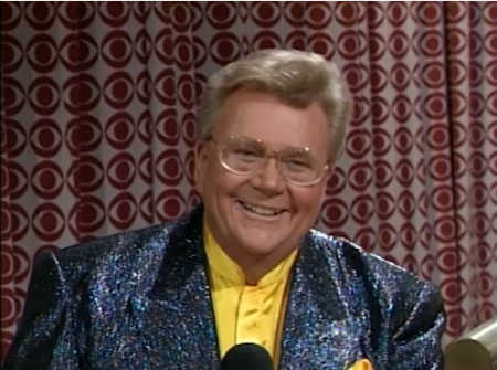 Rod is wearing a black/blue/purple sequined jacket & yellow collarless silk shirt with matching pocket square