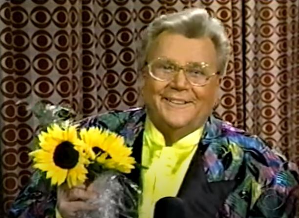 Rod is wearing a multi-color/patterned jacket, yellow collarless silk shirt and holding sunflowers