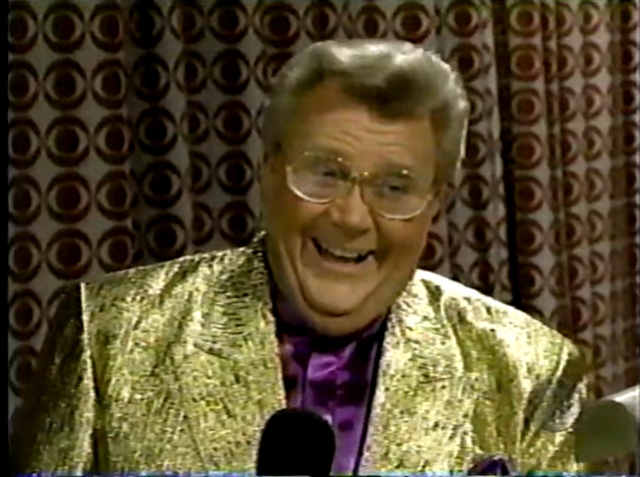 Rod is wearing a shiny gold jacket & purple collarless silk shirt with matching pocket square
