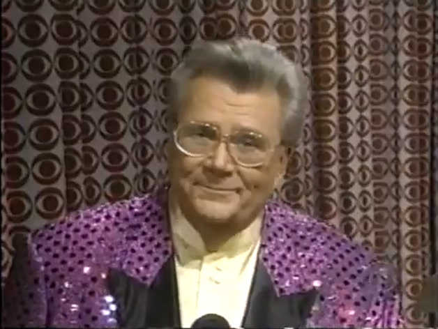 Rod is wearing a purple sequined with shiny black dots & black lapels jacket & a cream-colored collarless silk shirt