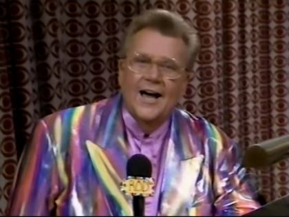Rod is wearing a shiny multi-colored jacket & violet collarless silk shirt with matching pocket square