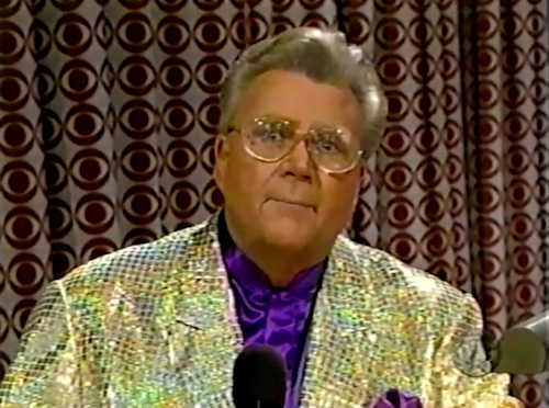 Rod is wearing a gold sequined jacket & purple collarless silk shirt with matching pocket square