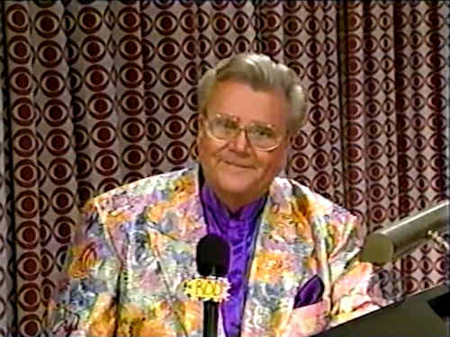 Rod is wearing a shiny multi-pastel-colored paisley-patterned jacket & purple collarless silk shirt with matching pocket square