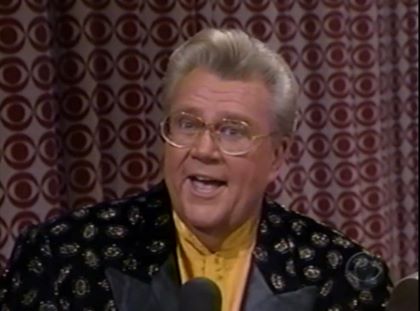 Rod is wearing a black jacket with silver splotches, black lapels & a mustard-yellow collarless silk shirt