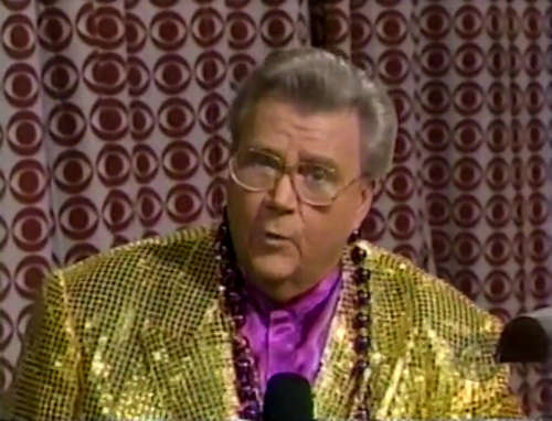 Rod is wearing a gold sequined jacket, purple collarless silk shirt & a string of gold & purple beads