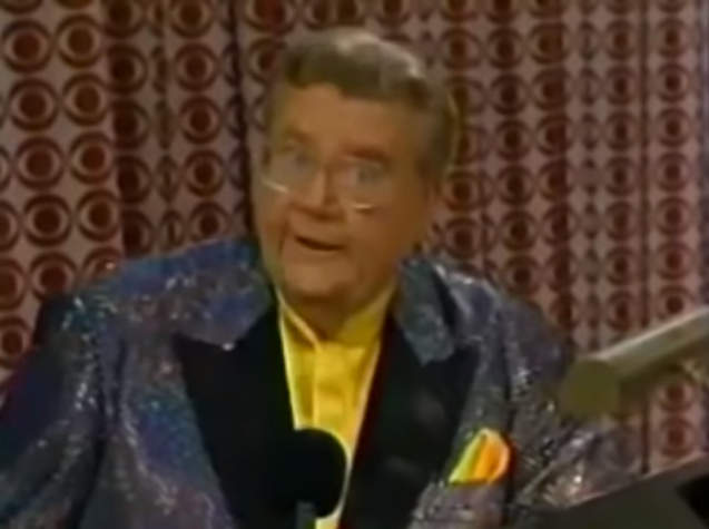 Rod is wearing a grey sequined jacket with black lapels, yellow collarless silk shirt with matching pocket square