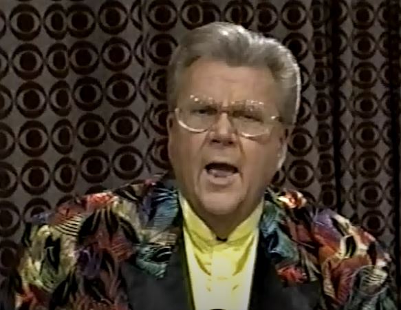 Rod is wearing a black jacket with a multicolored feather pattern, black lapels & yellow collarless silk shirt