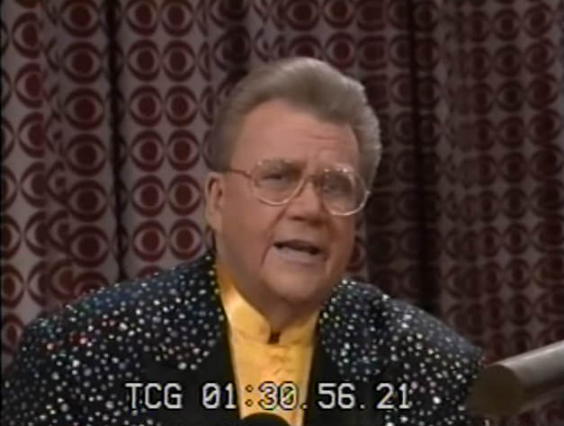 Rod is wearing a black jacket with white speckles, black lapels & mustard-yellow collarless silk shirt