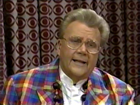 Rod is wearing a multi-colored gingham jacket, white collarless silk shirt & a beaded necklace