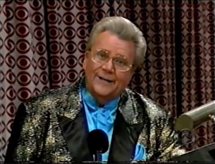Rod is wearing a shiny gold patterned jacket with black lapels & a light-blue collarless silk shirt with matching pocket square