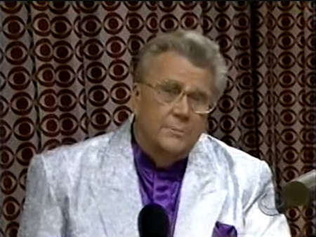 Rod is wearing a shiny silver jacket & purple collarless silk shirt with matching pocket square