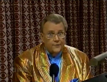 Rod is wearing a shiny brown/orange gingham jacket & blue collarless silk shirt with matching pocket square