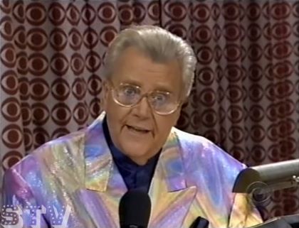 Rod is wearing a snihy violet/pink/yellow-fade jacket & dark-blue collarless silk shirt with matching pocketsquare