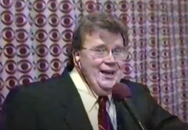 Rod is wearing a black suit coat, white shirt & red tie with thin white striping