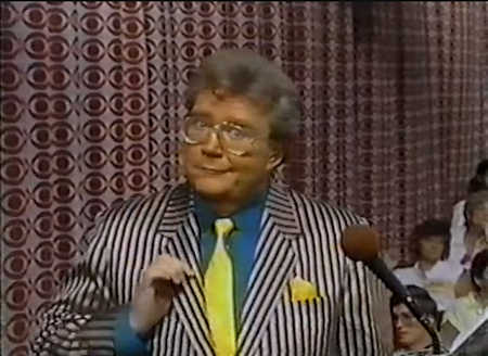 Rod is wearing a black/silver striped jacket, teal shirt and yellow necktie with matching pocketsquare