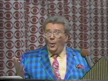 Rod is wearing a blue/dark blue gingham jacket, yellow shirt and a pink/red mottled tie with matching pocket square