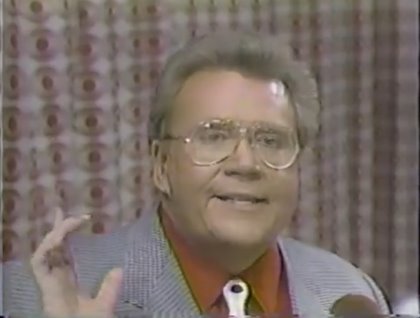 Rod is wearing a grey jacket, dark-orange shirt & a white neck-tie with polks dots