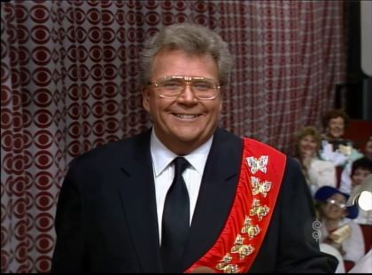 Rod is wearing a black suit, matching necktie, white shirt & red sash with gold butterflies