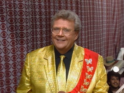 Rod is wearing a gold jacket, matching necktie, black shirt & red sash with gold butterflies