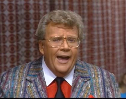 Rod is wearing a grey tweed jacket with purple/teal/red/white/yellow stitching, a black neck tie and a red shirt with white collars and matching pocketsquare