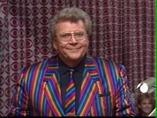 Rod is wearing a rainbow-striped jacket, matching necktie & black shirt