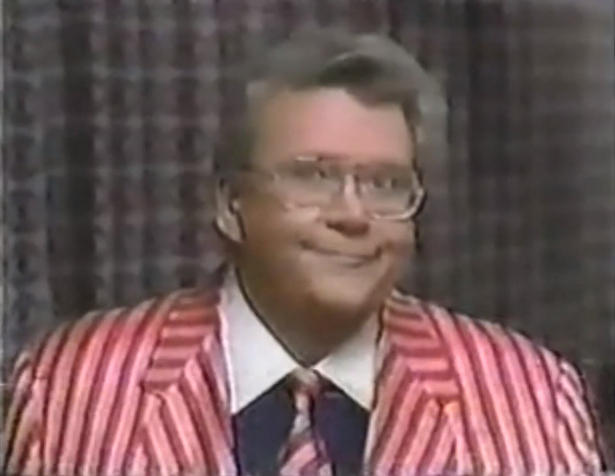 Rod is wearing a red/pink striped jacket with matching tie & a dark-blue shirt with a white collar