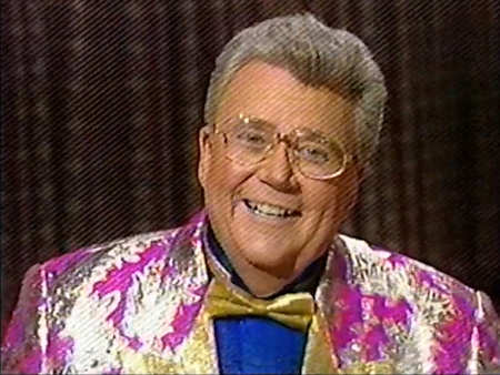 Rod is wearing a shiny fuchsia jacket with silver oak leaves & gold lapels, a gold bowtie & blue collarless silk shirt