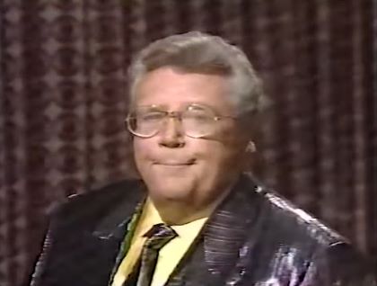 Rod is wearing a shiny black/black-striped jacket, matching necktie & yellow shirt