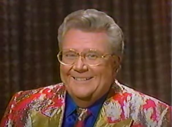 Rod is wearing a red jacket with a silver leaf pattern, gold lapels, matching necktie & blue silk shirt