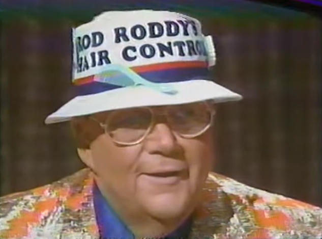 Rod is wearing an orange jacket with gold oak leaves, blue shirt & a bucket hat that reads Rod Roddy's Hair Control