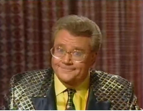 Rod is wearing a shiny gold jacket with a black-square pattern & black lapels, a matching tie & yellow shirt
