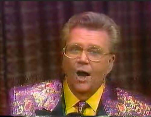 Rod is wearing a shiny gold & purple jacket with black lapels & matching tie with yellow shirt