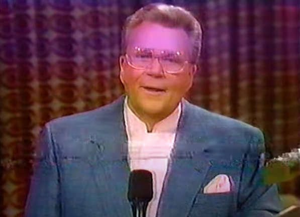 Rod is wearing a grayish-blue jacket, white pocket square & white collarless shirt