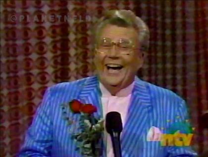 Rod is wearing a blue/blue striped jacket, pink collarless silk shirt, matching pocket square & is holding 2 red roses