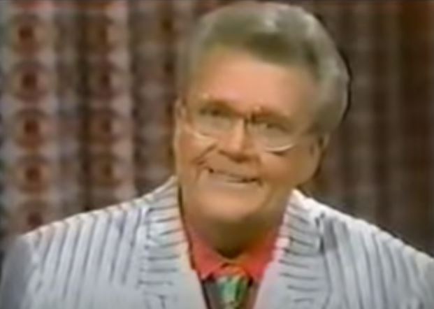 Rod is wearing a shiny white jacket with grey pinstripes, a salmon shirt & green necktie